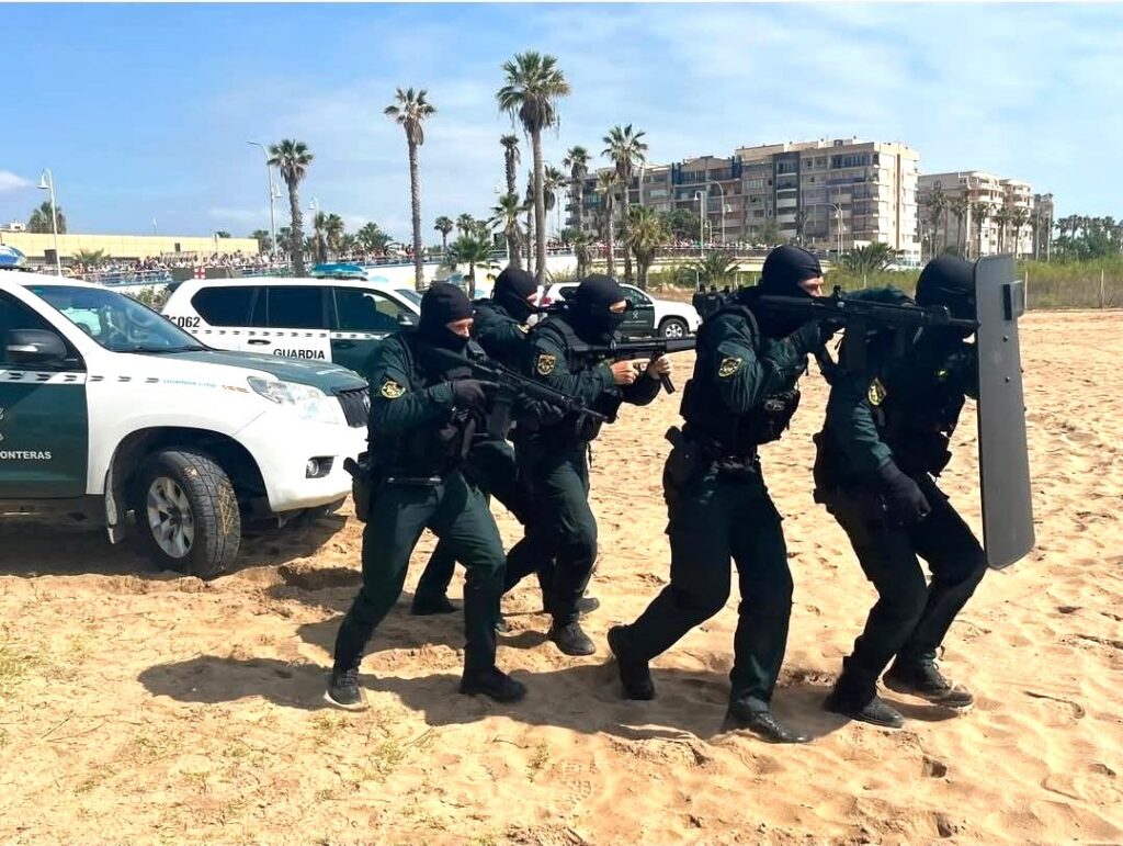 Tactical police unit in balaclavas advances on the sandy beach with rifles and a shielded formation, near patrol vehicles.