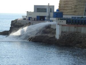 Industrial facility on a rocky coast releasing a jet of seawater into the sea from a large pipe.