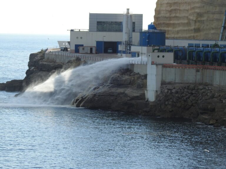 Industrial facility on a rocky coast releasing a jet of seawater into the sea from a large pipe.