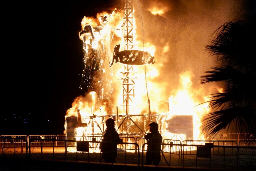 Nighttime scene of a tall wooden/steel framework burning, with bright flames and two silhouetted spectators behind barriers.