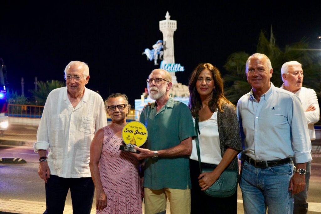 Six adults posing at night, holding a yellow 'Noche de San Juan 2026' award with a statue backdrop behind them.
