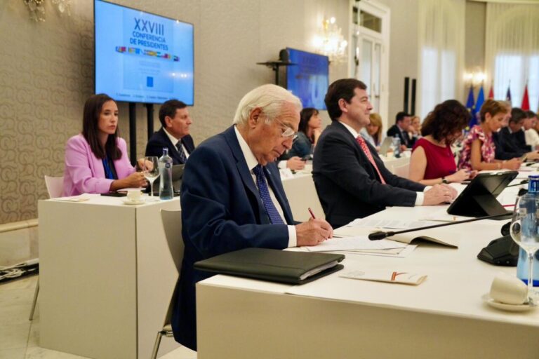 Senior man in a dark suit signs a document at a long white conference table during a formal meeting; colleagues sit and observe in a bright room with a screen and flags in the background.