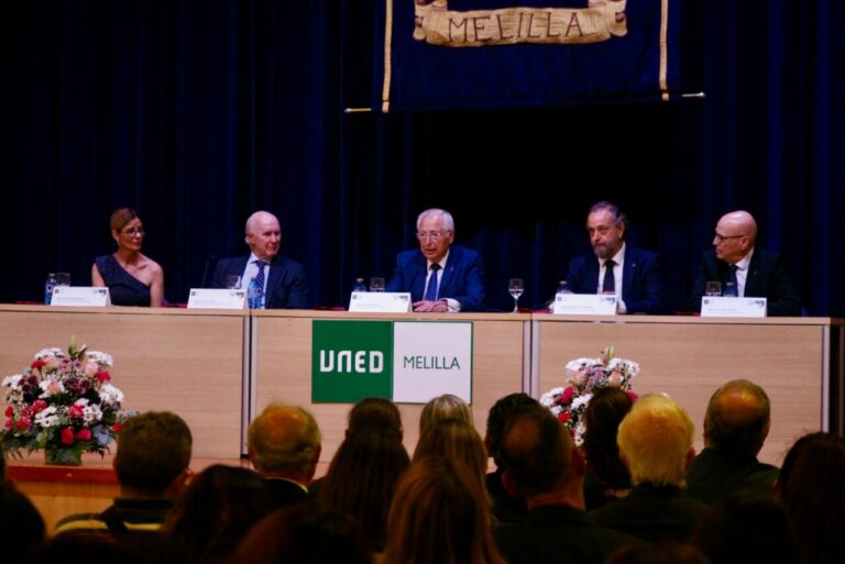 Panel of five people seated at a desk on a stage, with UNED Melilla banner and flower arrangements, addressing an audience