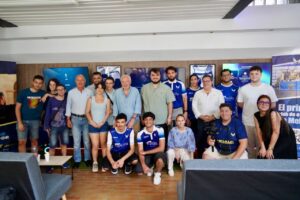 A large group photo of people indoors, some in blue team jerseys, posing together for a group shot near banners and screens in a lobby area.