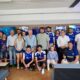 A large group photo of people indoors, some in blue team jerseys, posing together for a group shot near banners and screens in a lobby area.