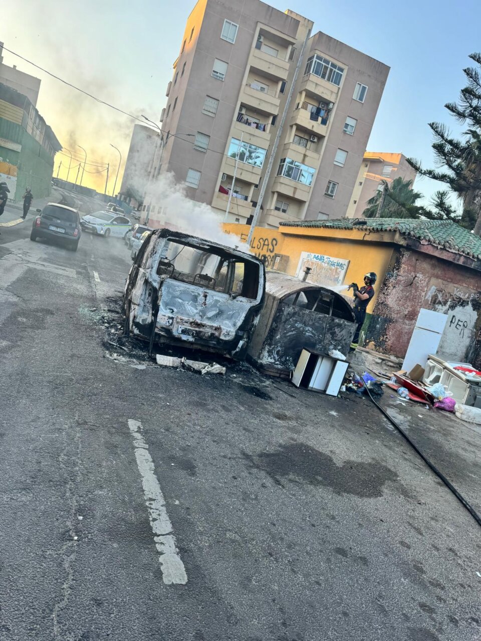 Charred van and car on a city street with smoke in the air and rubble scattered nearby, a person in protective gear nearby, with apartment buildings behind.