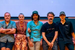 Five adults seated in a row, posing for a group photo against a blue backdrop on a stage. (Left to right: man in a teal shirt, woman in brown top with beads, man with a green cap, man in a black T‑shirt, young man in a cap)