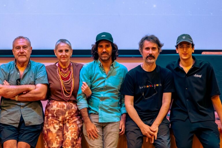 Five adults seated in a row, posing for a group photo against a blue backdrop on a stage. (Left to right: man in a teal shirt, woman in brown top with beads, man with a green cap, man in a black T‑shirt, young man in a cap)