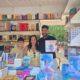 Family at an outdoor book stall with shelves of colorful books behind them and stacks of books on the table in front.