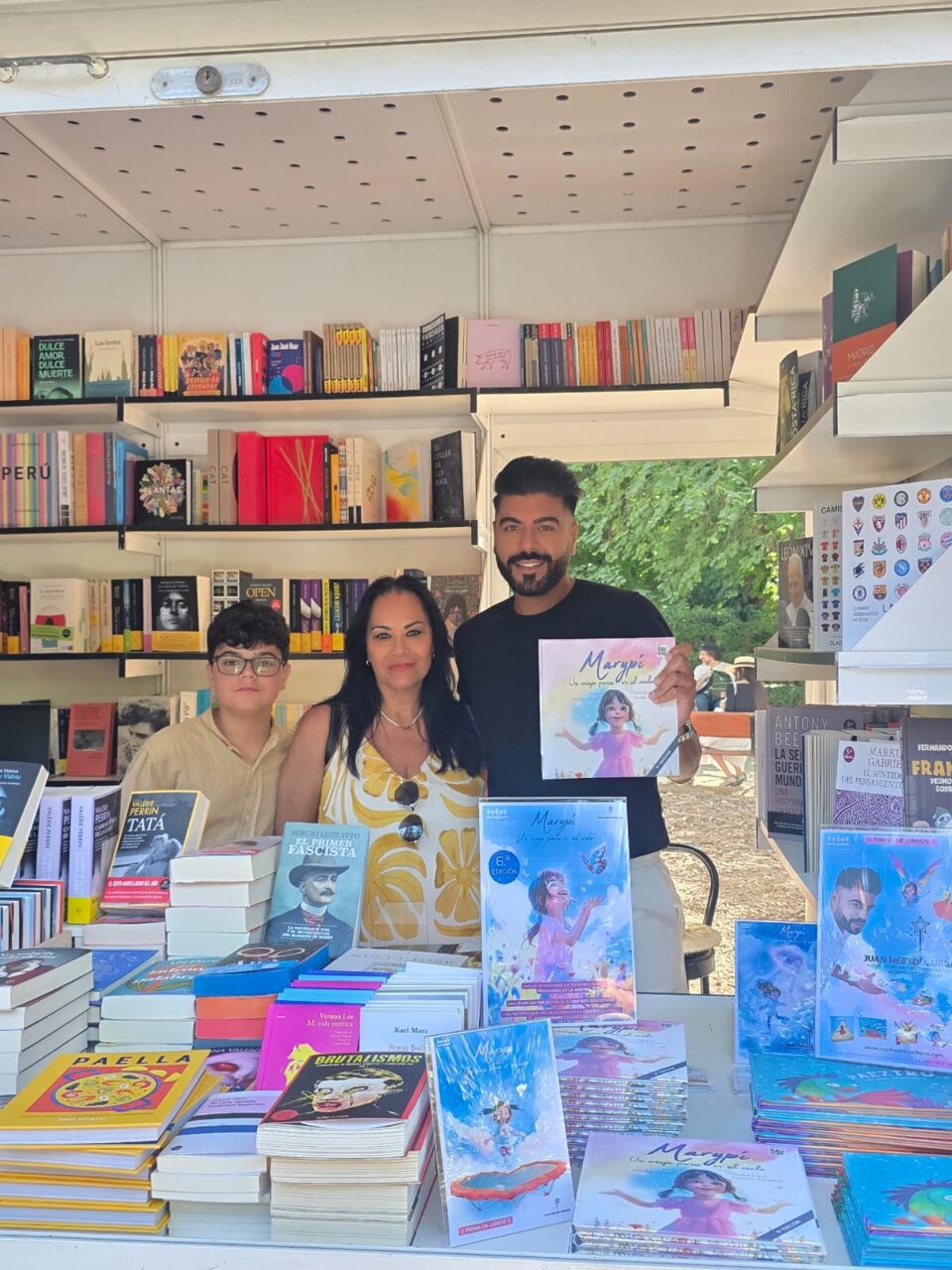 Family at an outdoor book stall with shelves of colorful books behind them and stacks of books on the table in front.