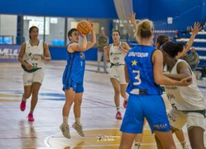 Female basketball player in blue jumps to shoot the ball while teammates and defenders watch on an indoor court.