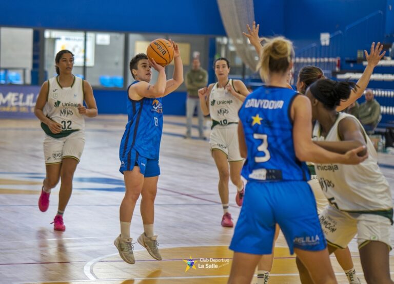 Female basketball player in blue jumps to shoot the ball while teammates and defenders watch on an indoor court.