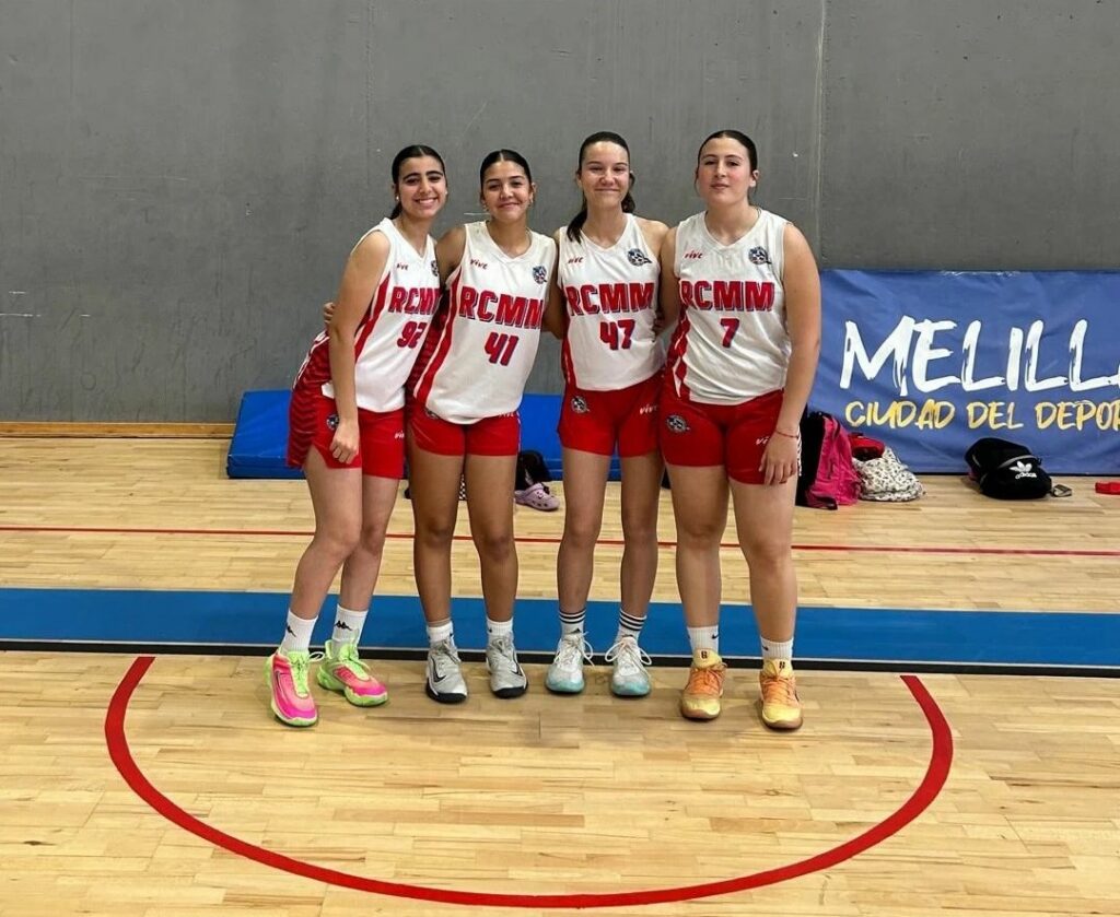 Four female basketball players in red-and-white uniforms (numbers 93, 41, 47, 7) pose together on a gym court, smiling.