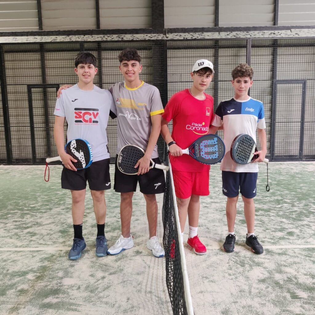 Four young men in sportswear stand side by side behind a padel net with their paddles, inside an indoor court with metal fencing in the background.