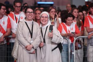 Two smiling nuns in beige robes with lanyards pose at a crowded event behind a metal barrier.