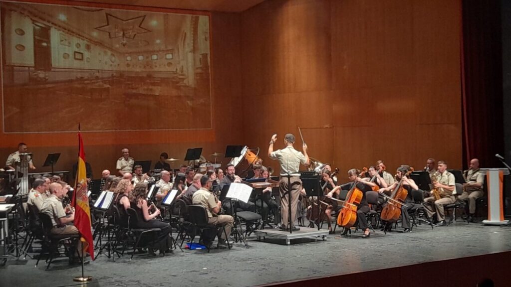 Military band on stage with conductor at center, musicians playing winds and strings; Spanish flag visible on the left.