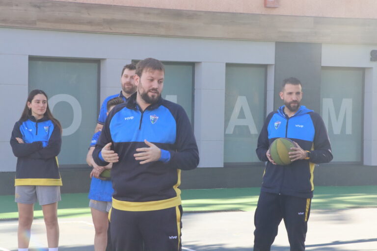 Coach in a blue-and-black tracksuit explains something to a group of teammates outside a modern building; a man on the right holds a basketball.