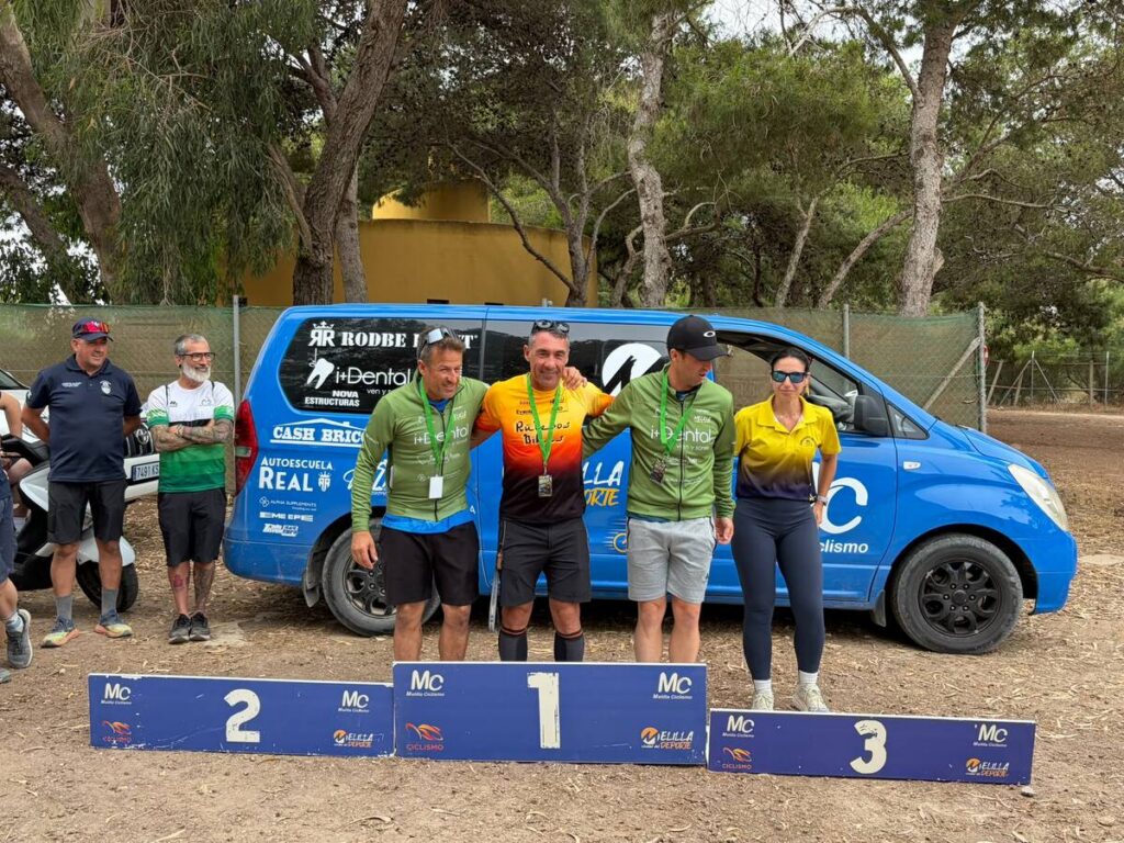 Three athletes on the first-place podium pose with medals, a blue sponsor van behind them and onlookers nearby.