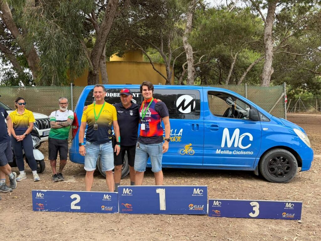 Group of cyclists with medals posing on a three-step podium in front of a blue Melilla Ciclismo van under trees.