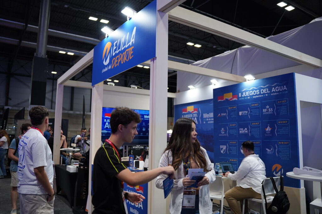 Trade show booth for Melilla Deporte with staff and visitors in a large exhibition hall.