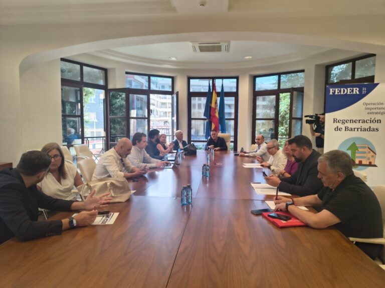 Group of people seated around a long conference table in a bright room; flags and a FEDER banner are visible in the background.