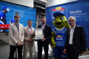 Five people in business attire pose with a green frog mascot at a trade show booth with a blue backdrop and screen behind them.