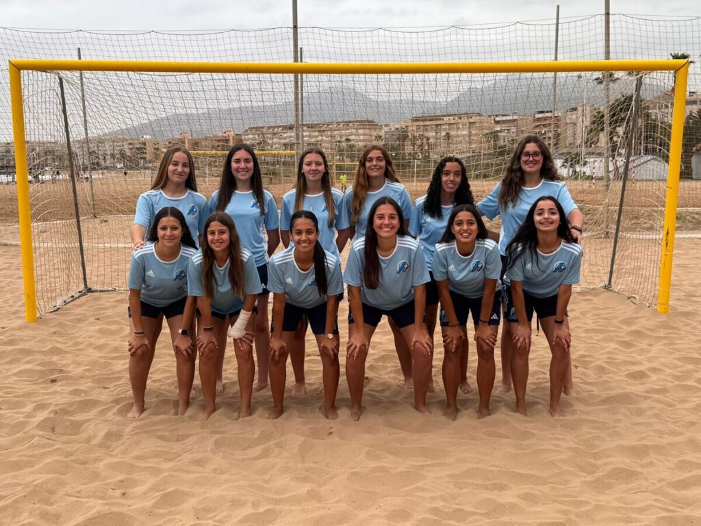 Twelve girls in light blue jerseys pose for a team photo in front of a yellow goal on a sandy beach soccer field, smiling at the camera.