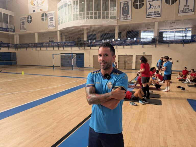 Man in a blue athletic shirt stands with arms crossed in a gymnasium, a fitness class with participants on mats visible in the background.