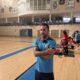 Man in a blue athletic shirt stands with arms crossed in a gymnasium, a fitness class with participants on mats visible in the background.