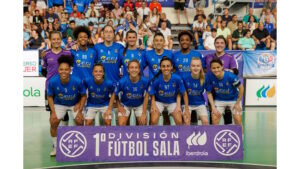 A women's futsal team in blue jerseys kneels and stands together for a group photo in an indoor arena, with spectators in the stands behind them and a purple banner reading '1ª División Fútbol Sala' in front.