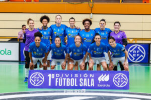 Women’s futsal team posing for a group photo in blue jerseys on an indoor court, behind a purple banner that reads 1ª División Fútbol Sala.