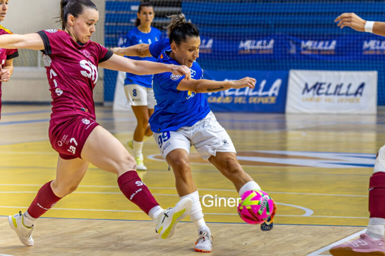 Two female players in blue and maroon jerseys contest a ball on an indoor futsal court, emphasizing action and competition.