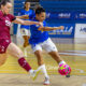 Two female players in blue and maroon jerseys contest a ball on an indoor futsal court, emphasizing action and competition.