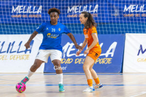 Two female futsal players contesting the ball on an indoor court; blue jersey controls the ball while orange jersey defends near the goal area.