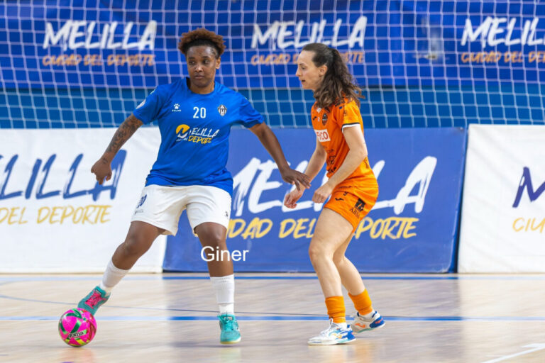 Two female futsal players contesting the ball on an indoor court; blue jersey controls the ball while orange jersey defends near the goal area.