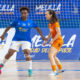 Two female futsal players contesting the ball on an indoor court; blue jersey controls the ball while orange jersey defends near the goal area.