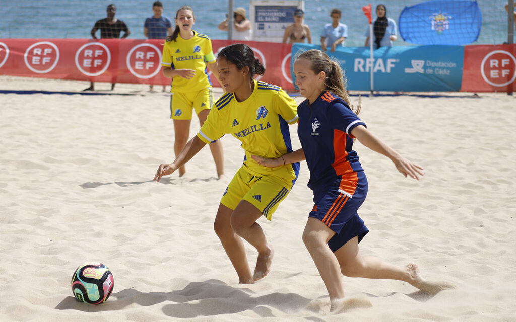 Two girls in yellow and blue uniforms chase a colorful soccer ball on a sandy beach during a match, with spectators and banners in the background.