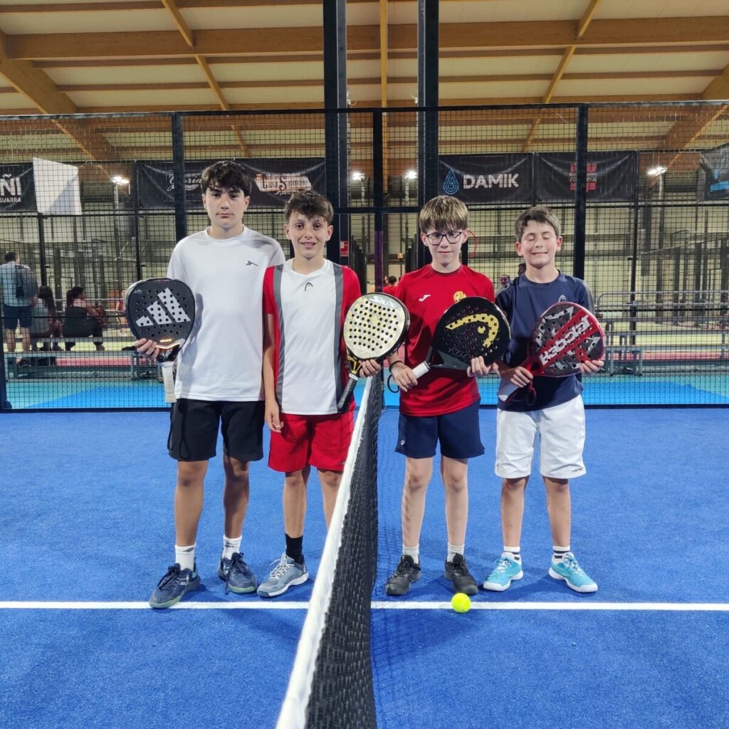 Four boys stand side by side on a blue indoor padel court, each holding a paddle.