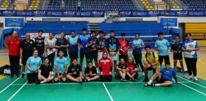 Group of young athletes on a badminton court posing with trophies in a sports hall, blue and yellow bleachers behind.