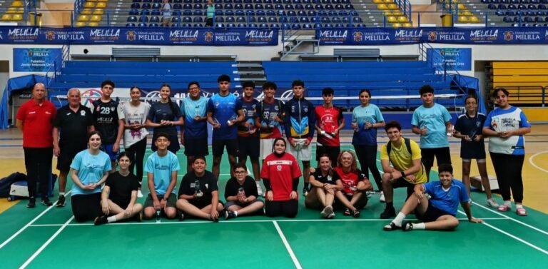 Group of young athletes on a badminton court posing with trophies in a sports hall, blue and yellow bleachers behind.