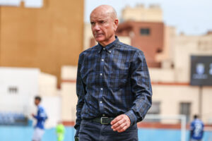 Older man with a shaved head in a blue plaid shirt walking on an outdoor sports field with blurred players in the background at a stadium."