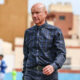 Older man with a shaved head in a blue plaid shirt walking on an outdoor sports field with blurred players in the background at a stadium."