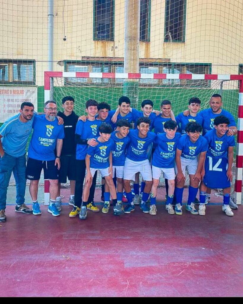 Group of young soccer players in blue jerseys and white shorts posing together in front of a goal on an indoor court, with two adults on the sides.