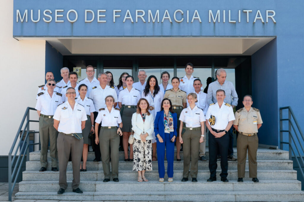 Group portrait of military officers and civilians standing on the entrance steps of the Museo de Farmacia Militar, a blue building with the sign above.