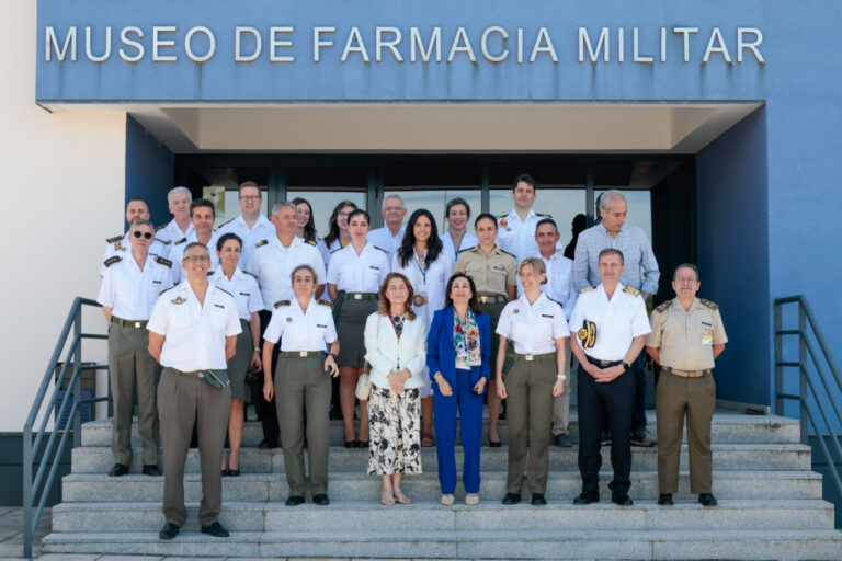 Group portrait of military officers and civilians standing on the entrance steps of the Museo de Farmacia Militar, a blue building with the sign above.
