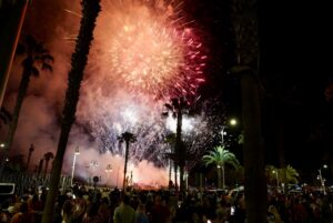 Crowd watches bright fireworks over palm trees at night, with smoke and streetlights.