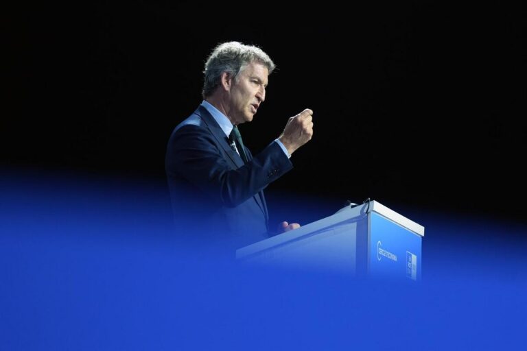 Man in a dark blue suit giving a speech at a podium on a dark stage, with a blue foreground blur partially obscuring the view.