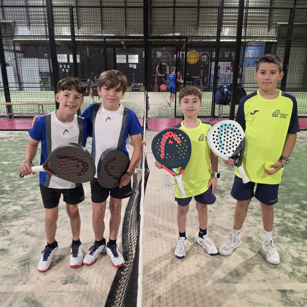 Four young boys stand at a padel court net, wearing blue/white and neon shirts, each holding a padel racket and a ball nearby in a group pose.