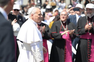 A man in white papal robes walks past clergy in black with magenta sashes as a crowd applauds in the background.