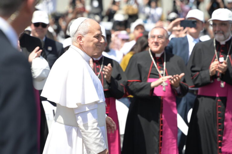 A man in white papal robes walks past clergy in black with magenta sashes as a crowd applauds in the background.
